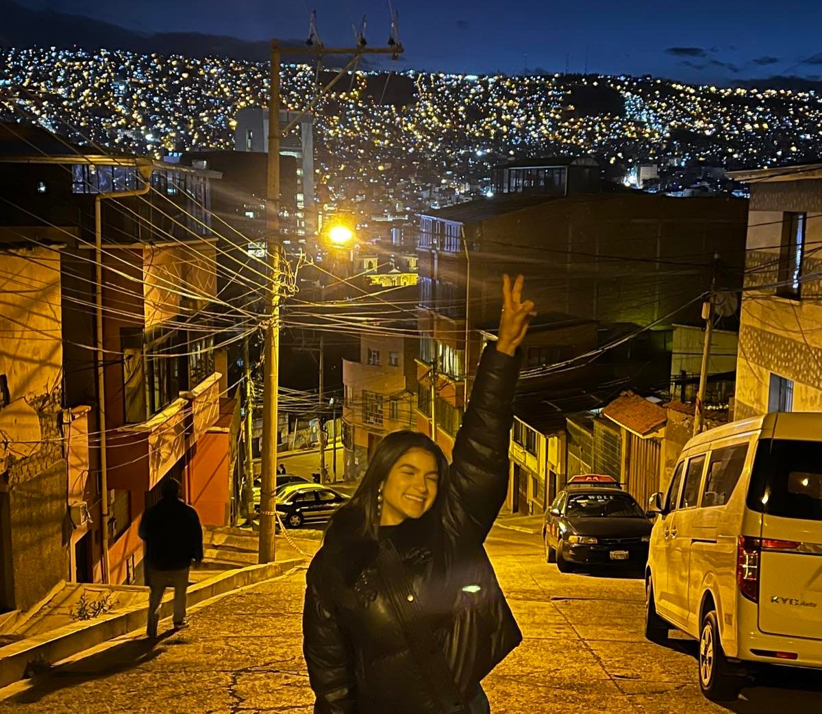 Mujer en una calle empinada de La Paz de noche, con vehículos y las laderas iluminadas al fondo