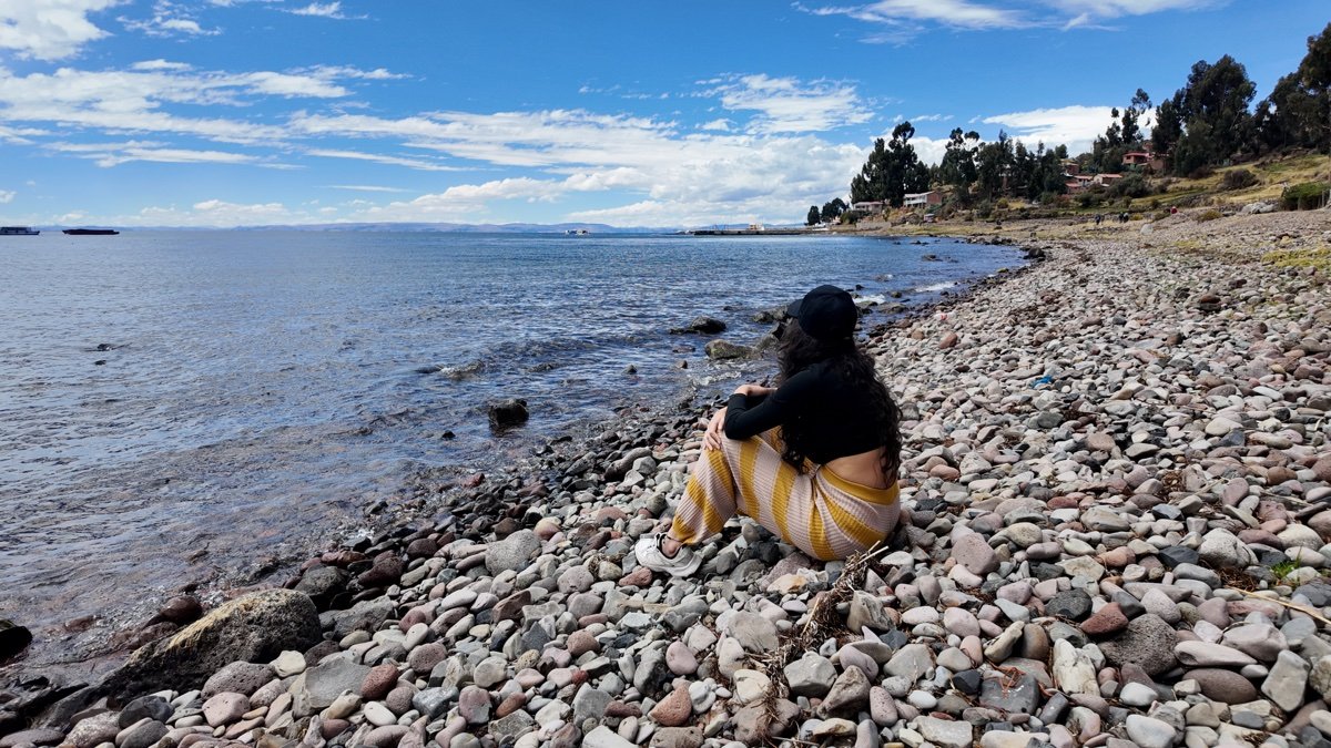Mujer sentada en una orilla de piedras del lago Titicaca, con árboles y casas al fondo